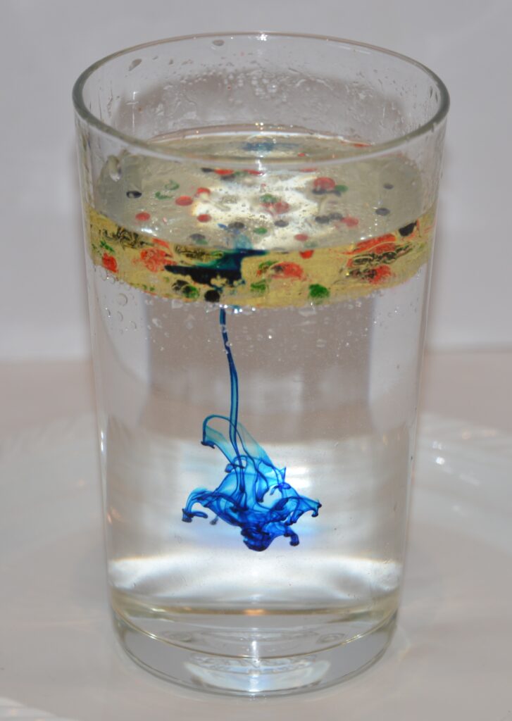 Vaso alto lleno de agua y una capa de aceite de cocina. Remolinos de colorante alimentario se disparan a través del aceite en el agua.