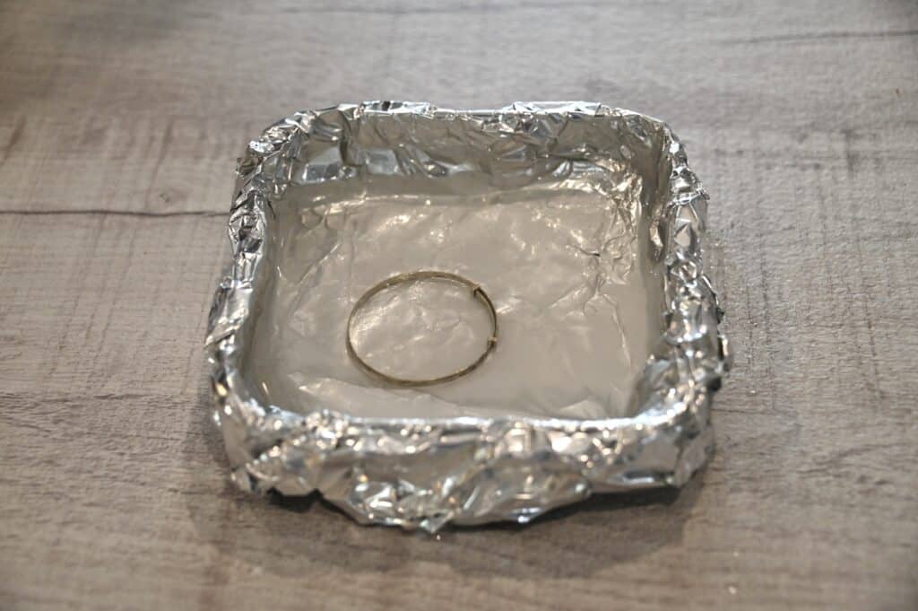 Plastic container lined with aluminum foil. Inside the container is a silver bracelet covered with boiling water and baking soda.